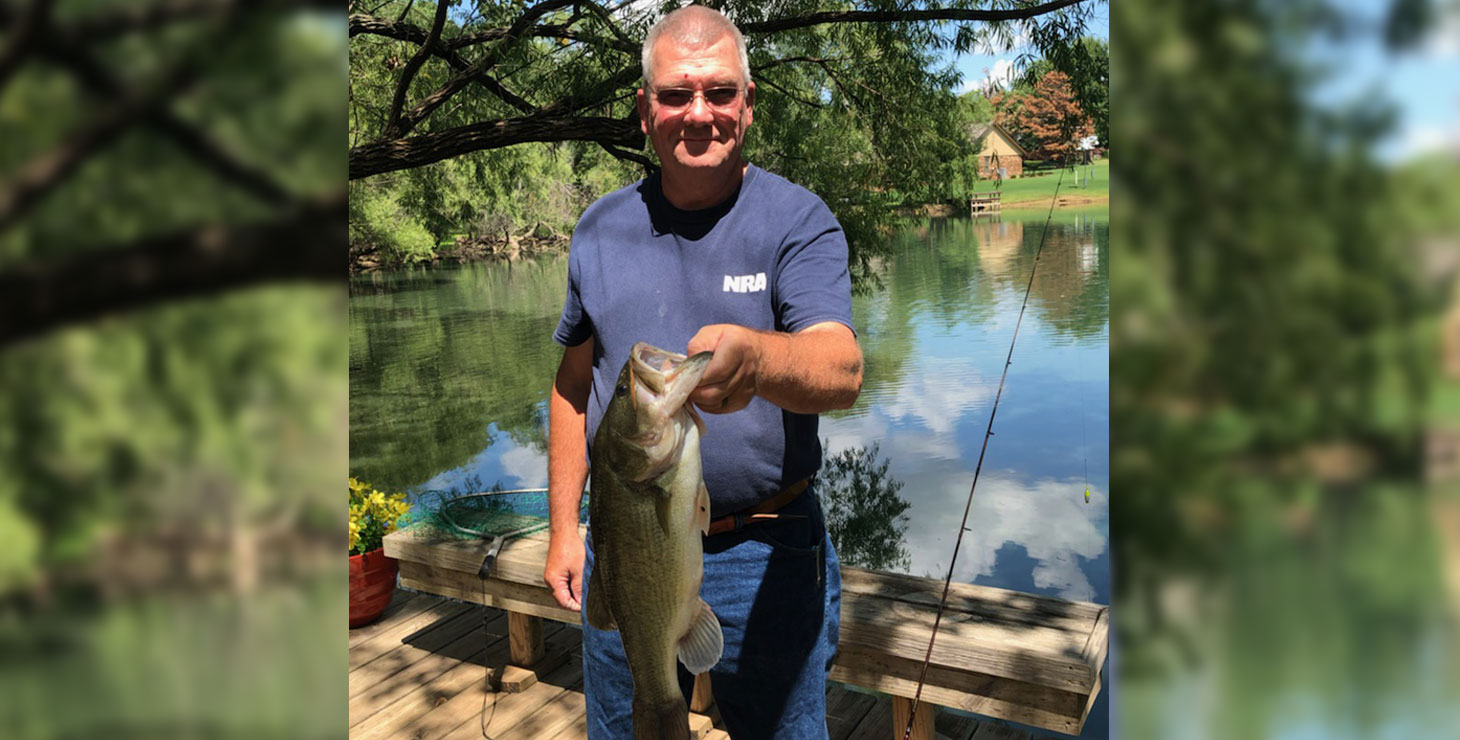 Jerry Kates is wearing a navy t-shirt and holding a bass fish. 